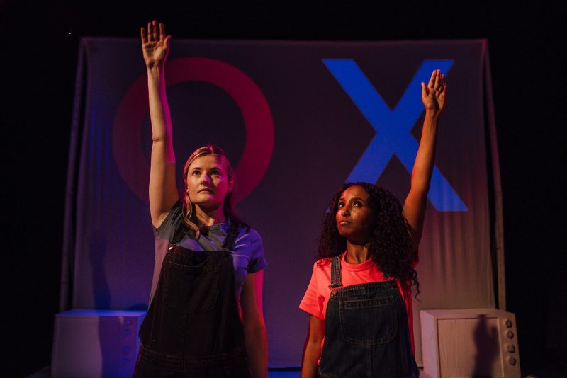 On stage photo of two women stood up with one hand raised in the air: one woman looks confident and the other more uncertain