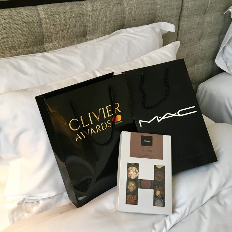 olivier awards goodie bag, mac cosmetics goodie bag, and hotel chocolat box of confectionary arranged on top of a made bed