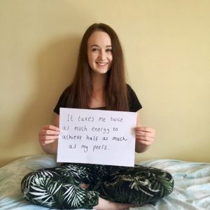 pippa sat crossed legs on bed with a4 sign reading 'it takes me twice as much energy to achieve half as much as my peers'