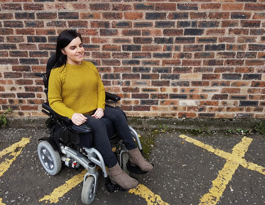 emma in powerchair wearing mustard knitted jumper, jeans and boots, in front of brick wall background