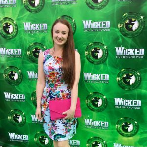 pippa standing in front of green wicked backdrop, wearing floral dress with pink clutch bag