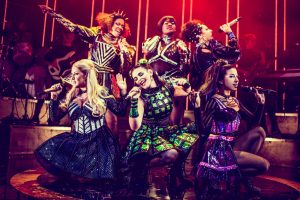 six queens posed together on stage with microphones