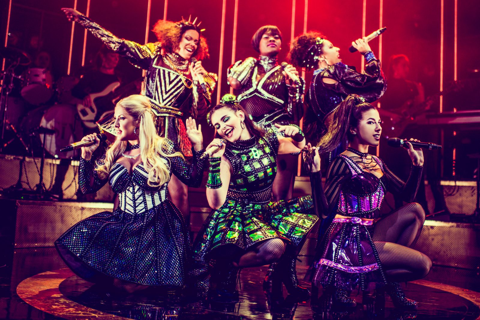 six queens posed together on stage with microphones