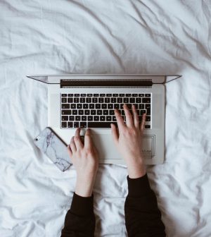 image taken from above of silver laptop open on white bedsheets with hands typing on keyboard, iphone laid on the left hand side