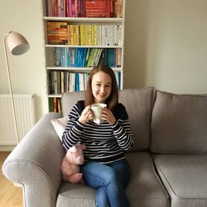 pippa sat on grey sofa holding cup of tea and smiling, with rainbow bookshelves in background