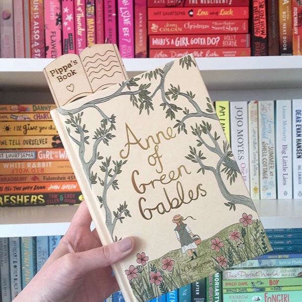 hand holding beige illustrated Anne Of Green Gables cover up in front of rainbow bookshelves
