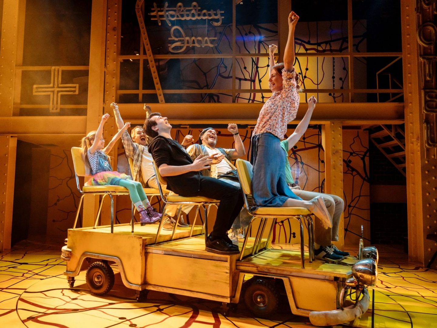 Cast of Little Miss Sunshine on stage sitting in yellow seats representing campervan, laughing and cheering