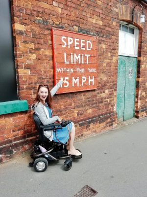 pippa in power-chair, pointing to outdoor sign reading 'speed limit within this yard 15MPH' and laughing