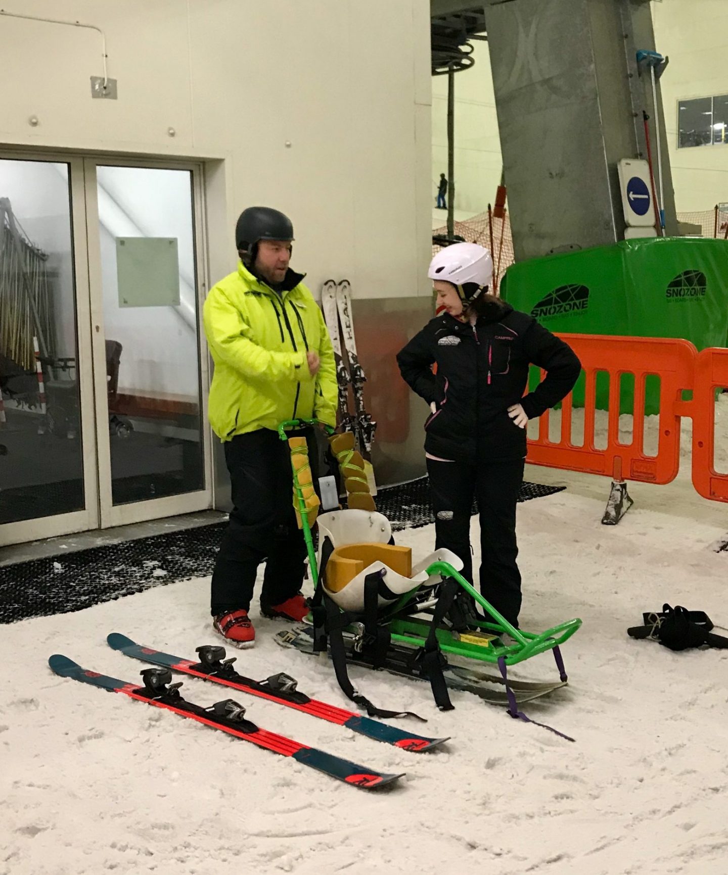 pippa and coach adam standing up and talking on the edge of the slopes, with sit-ski placed in front of them