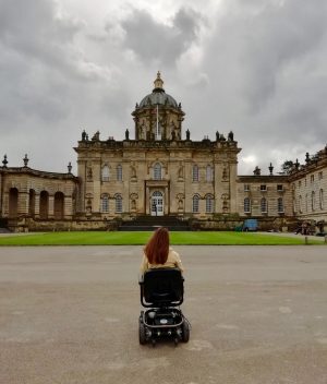pippa in powerchair with back to camera, with castle howard stately house in front
