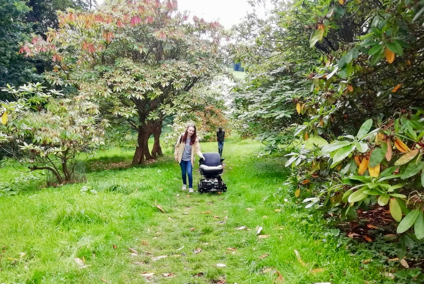 pippa walking alongside and driving powerchair up grassy hill, facing camera
