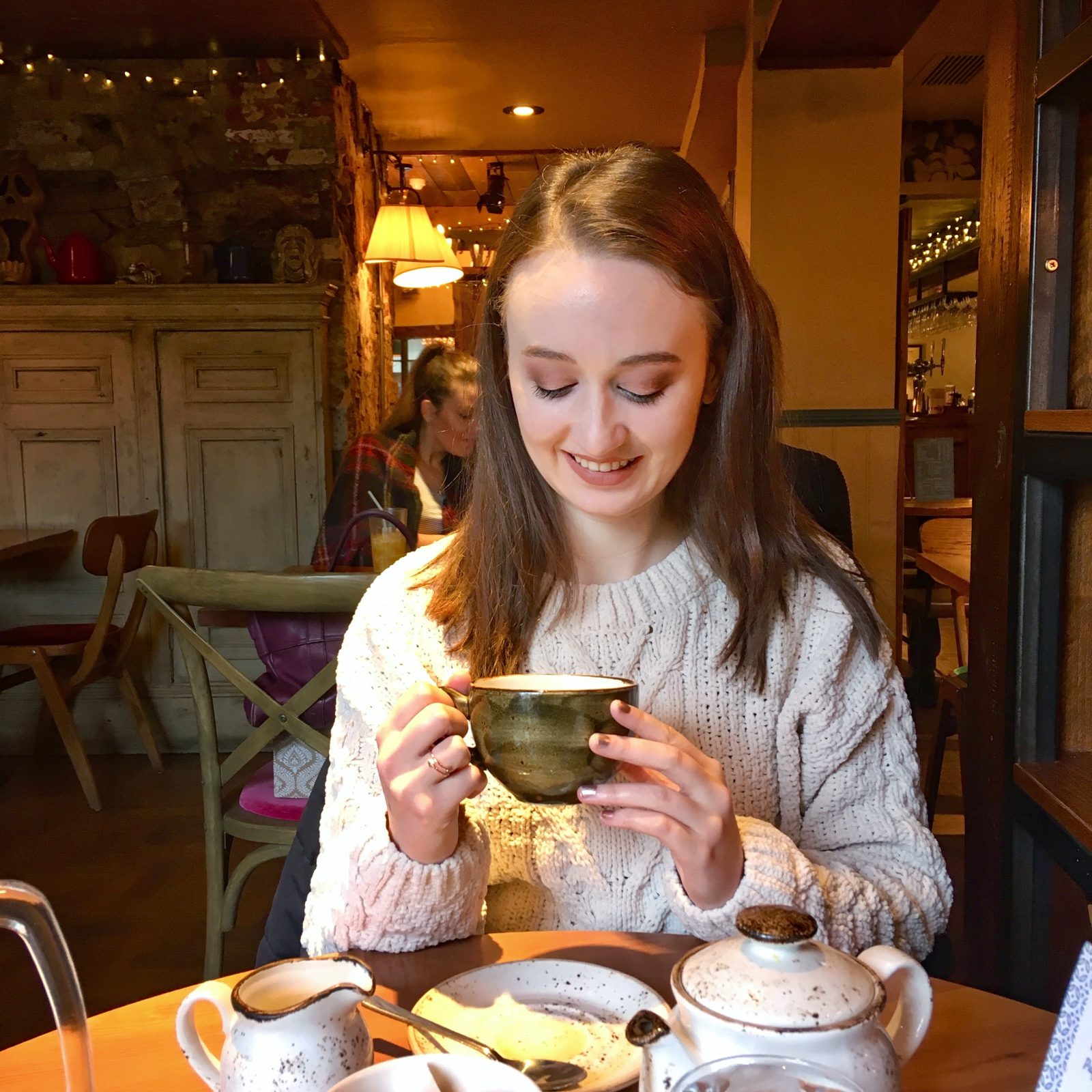 pippa sat at table wearing jumper and looking down at tea cup in hands, with tea pot on table in front