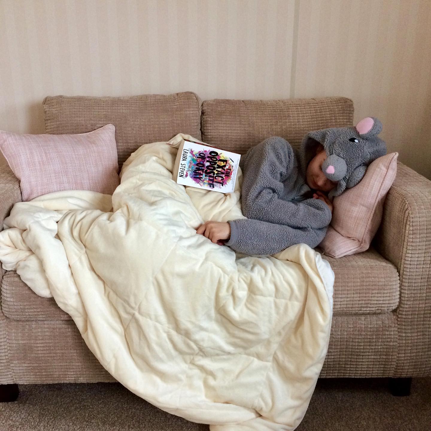 pippa laid on sofa in mouse onesie, under duvet with open book resting on top