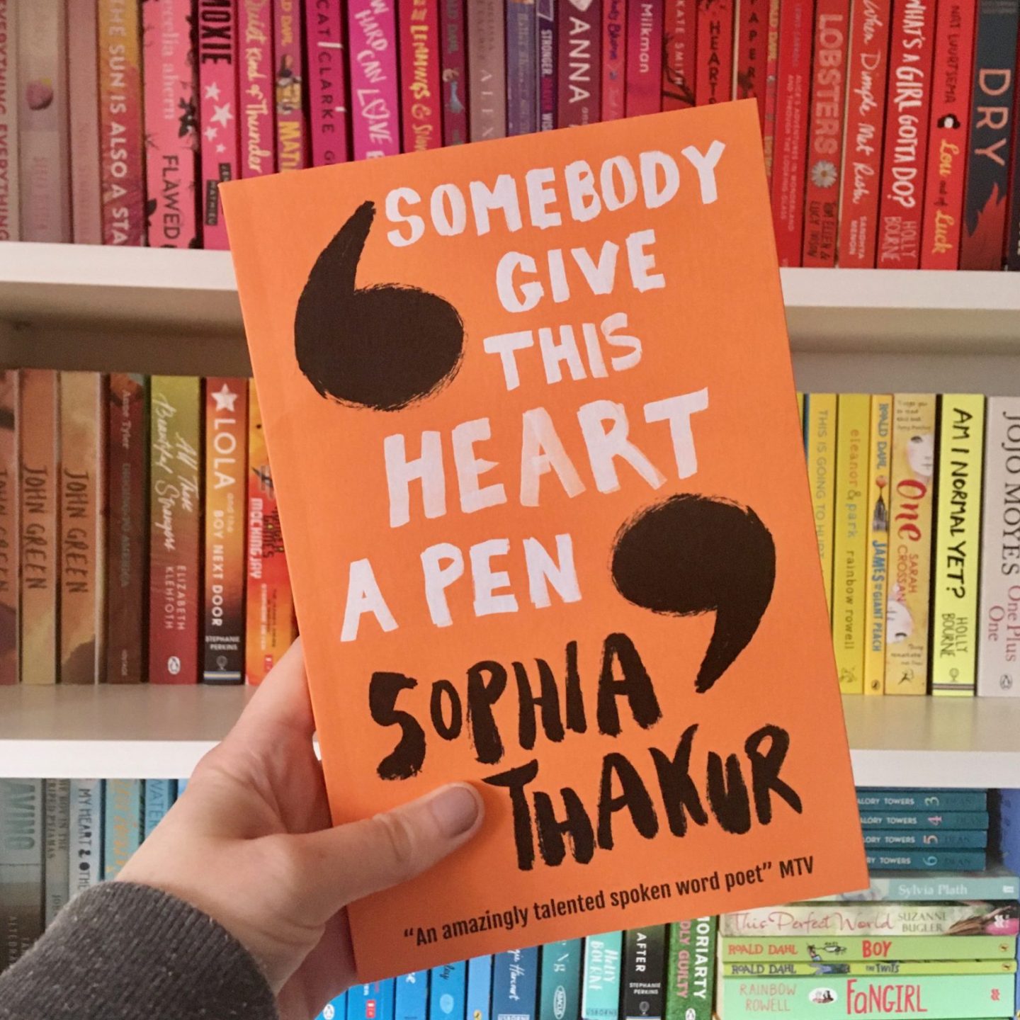 hand holding orange 'somebody give this heart a pen' book in front of rainbow bookshelves