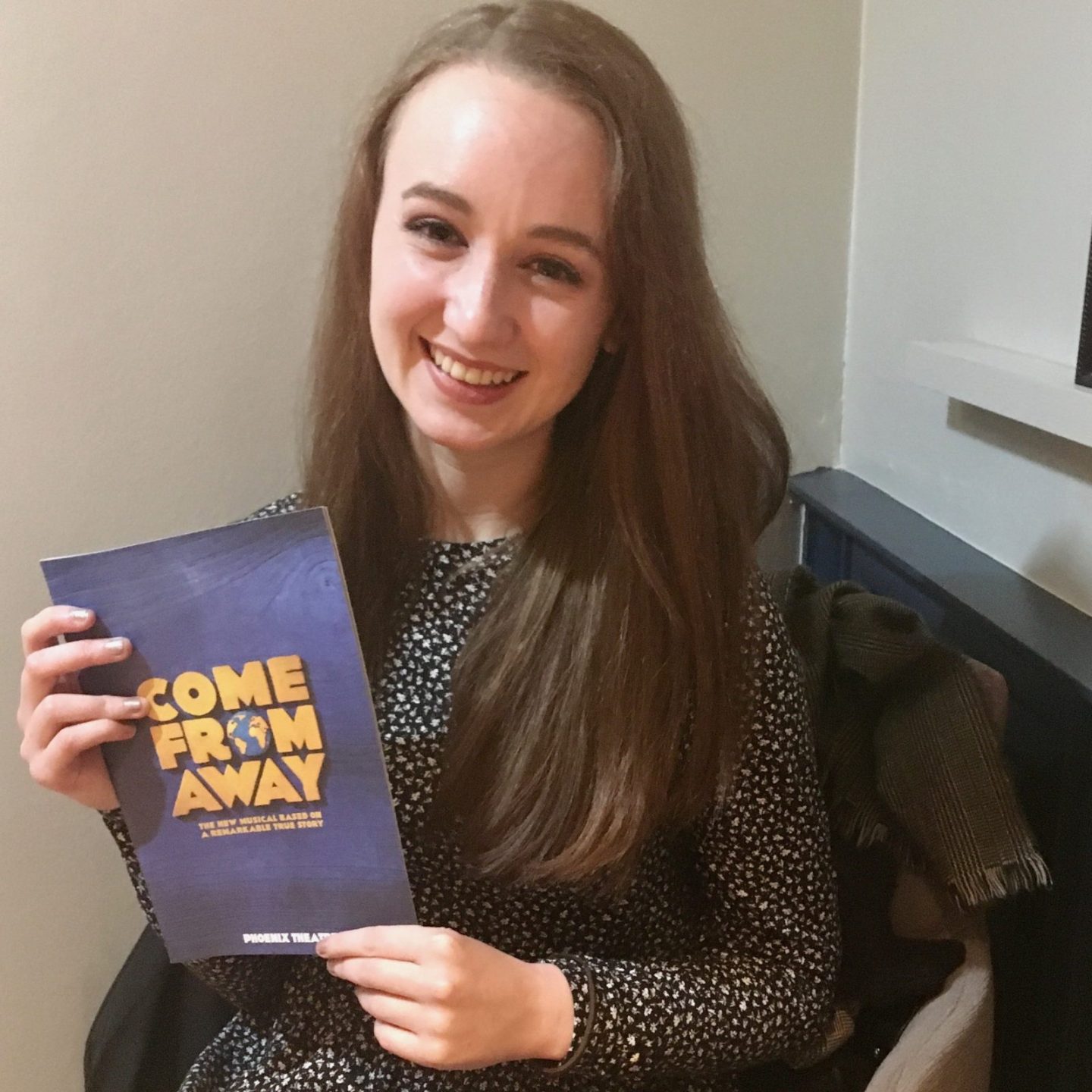 pippa sat on chair, holding up blue come from away programme and smiling