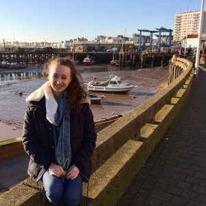 pippa sat outdoors wearing coat and scarf, harbour visible in background