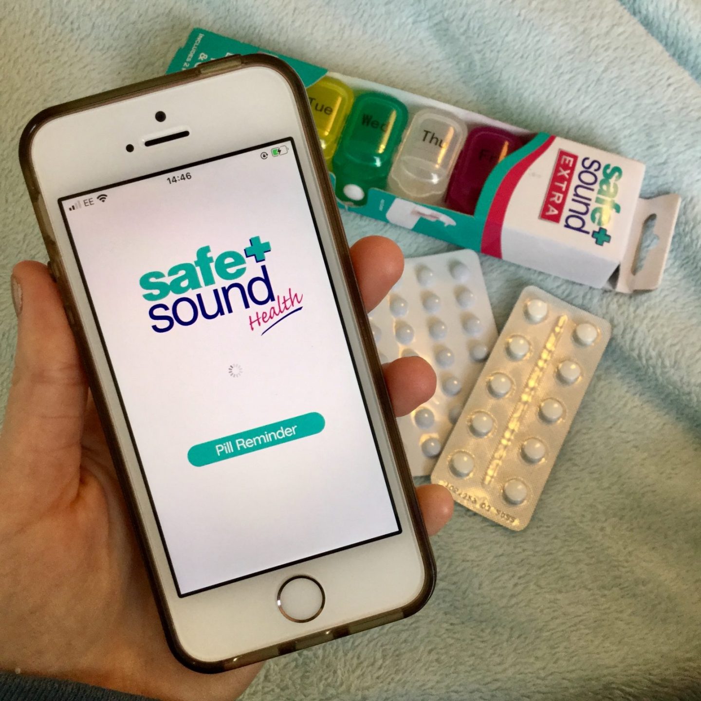 pippa's hand holding iphone displaying safe and sound medication reminder app, with pill box and tablets visible in the background