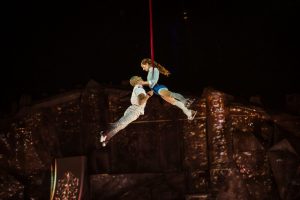 male and female performer hanging onto each other with legs raised, suspended in the air by red silk