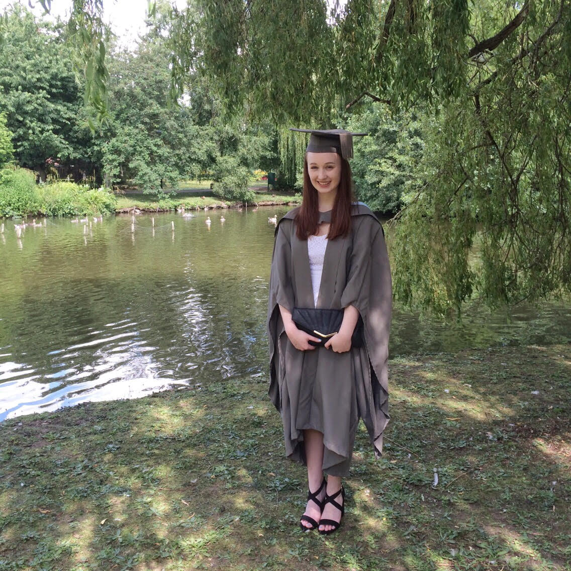 pippa wearing white dress with graduation cap and gown in grey, stood up outdoors with trees and river in background, holding black clutch bag and smiling