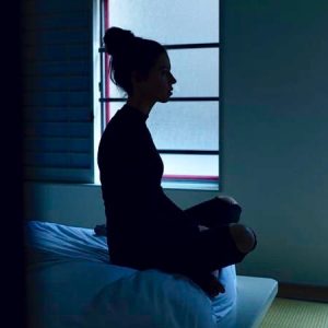 silhouette of woman sitting cross legged on bed, facing sideways, in front of a closed window