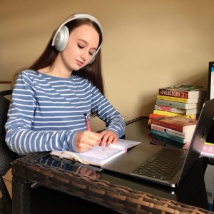 pippa sat at desk wearing stripy shirt and silver noise cancelling headphones, writing in a diary with open laptop in front of her and books in the background