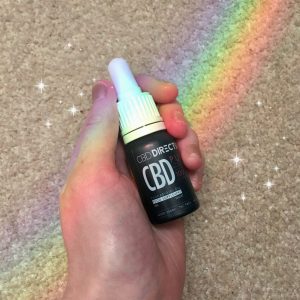 pippa's hand holding small black and white bottle of '10% cbd oil' in mixed berry flavour by CBD direct to you. Rainbow effect in background and sparkling star special effects.