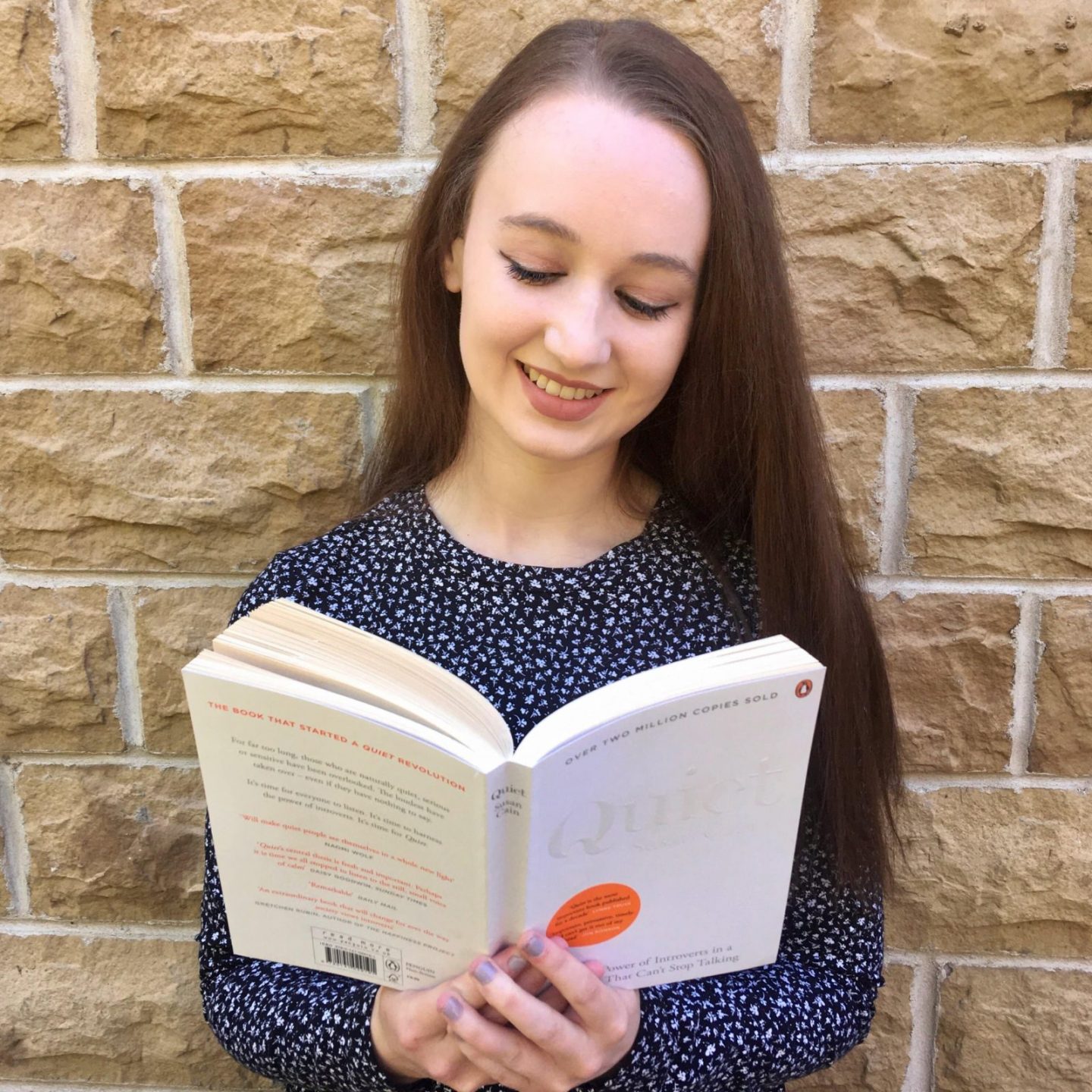 headshot of pippa outdoors in front of brick wall, hair down and wearing dark blue spotted dress, looking down at open book and smiling. book is 'quiet' by susan cain, plain white cover
