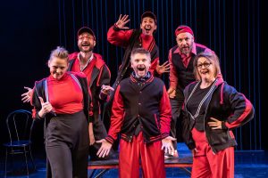 seven members of showstopper cast dressed in red and black, mid-performance on stage