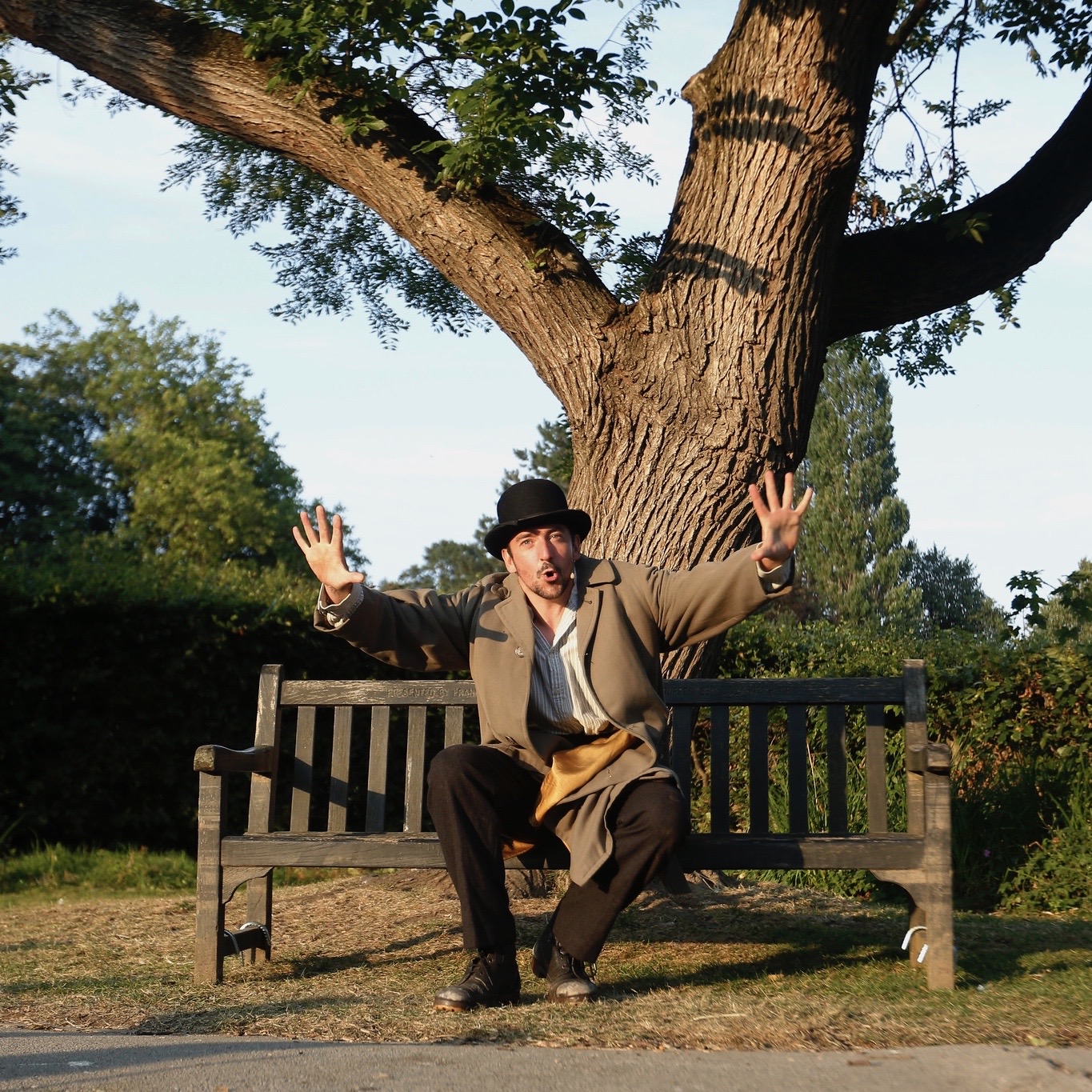 actor chris hannon in tan overcoat and black hat, sat on park bench in front of tree, gesturing with arms wide out and fingers splayed. image credits: Northedge Photography