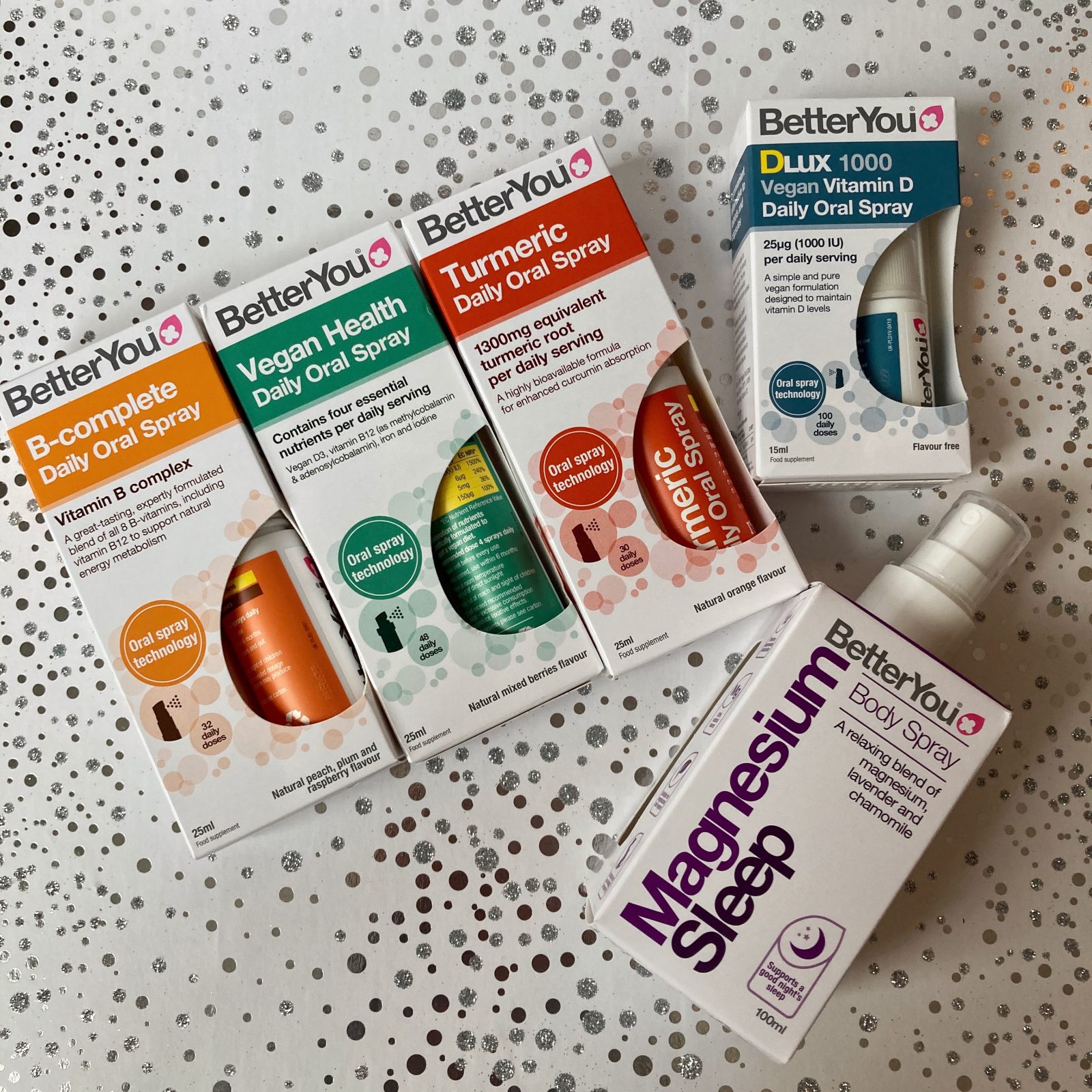 four of BetterYou's colourful transdermal sprays, turmeric, vegan health, b-complete and vitamin d, and magnesium sleep spray, flatlayed on top of white and silver sparkly background