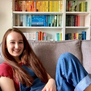 Pippa comfortably propped up on grey sofa, one leg bent up. Hair down, wearing red t-shirt and blue demin dungarees. Shelves of rainbow bookshelf (mostly blue and yellow) visible in background.