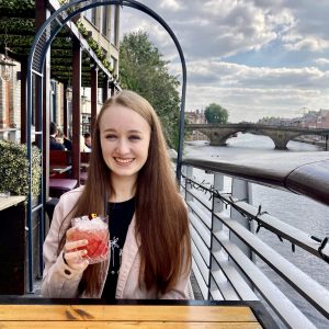 pippa sat at table outdoors with river visible in background. pippa is wearing a pink denim jacket with a black top under it, holding up a red 'mocktail' and smiling