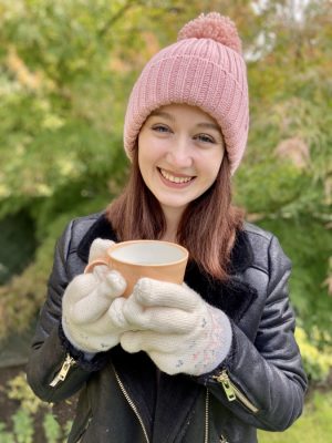 portrait image of pippa outdoors, holding a mug and smiling at camera. pippa is wearing pale pink bobble hat, black leather jacket and chunky cream coloured thermal gloves from heat holders