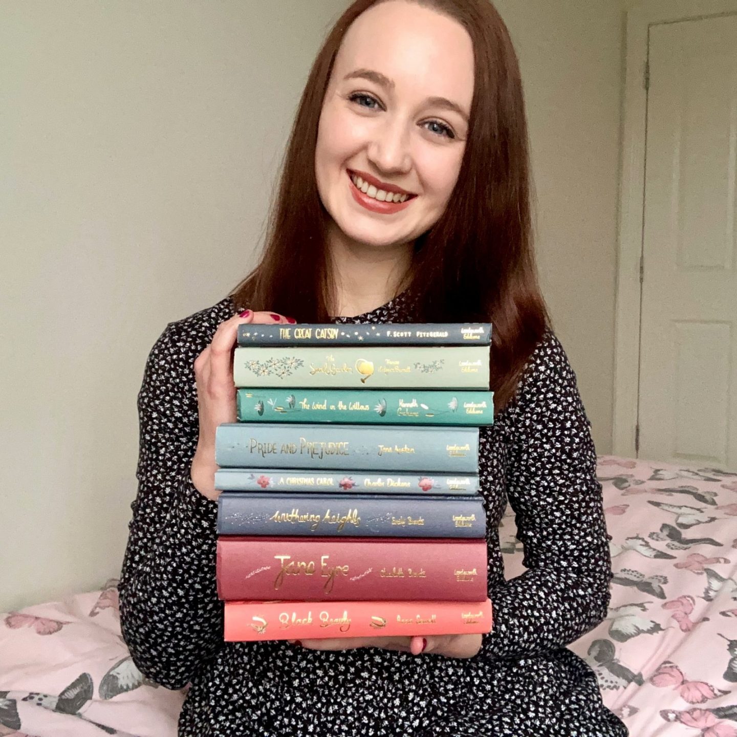 Pippa sat on edge of bed, holding up a huge pile of books and smiling. Pippa is wearing long-sleeved navy blue dress with tiny white spots, with brown hair down. Books are all classics from Wordsworth Collector’s Editions in a rainbow spectrum of pastel colours and titles embroidered in gold. Books top to bottom are The Great Gatsby, The Secret Garden, The Wind In The Willows, Pride and prejudice, a Christmas Carol, withering heights, Jane Eyre, and black beauty.￼￼
