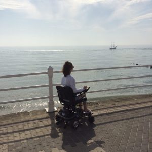 pippa using small black power-chair along the seafront, pippa turned away and looking out to sea which is visible in background