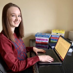 Pippa sat at desk working on laptop, hands on keyboard and looking down at screen. Pippa has hair down and is wearing a burgandy woollen jumper, piles of paperback books in background