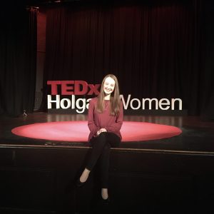pippa sat on edge of stage at TEDx, iconic red carpet and lettering in background. Pippa is wearing a burgandy long sleeved top, black jeans and black pumps.
