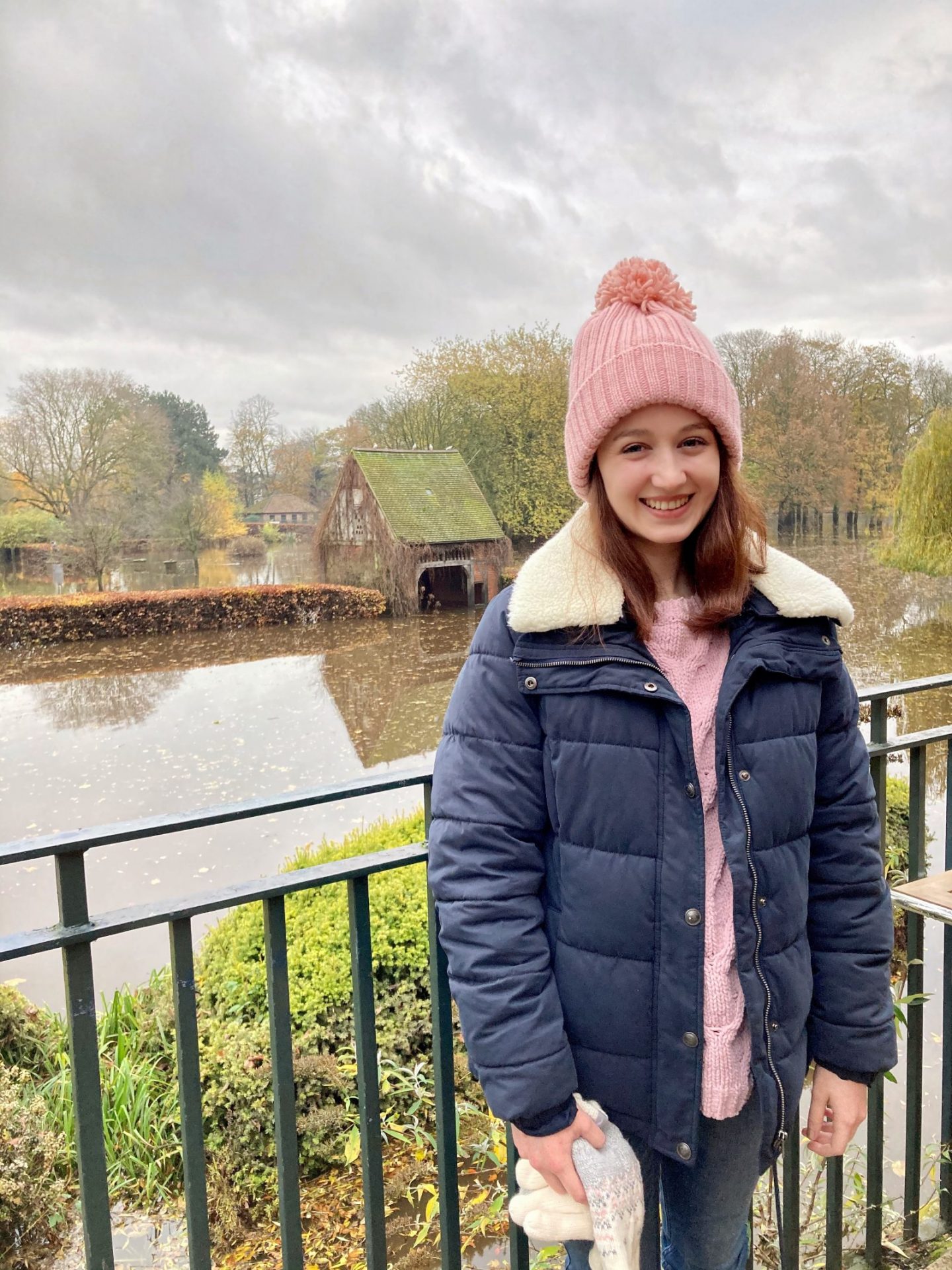 Pippa stood outdoors and smiling, wearing navy blue winter coat, lilac jumper and pale pink bobble hat. Park visible in back (rather flooded with water at the time!).