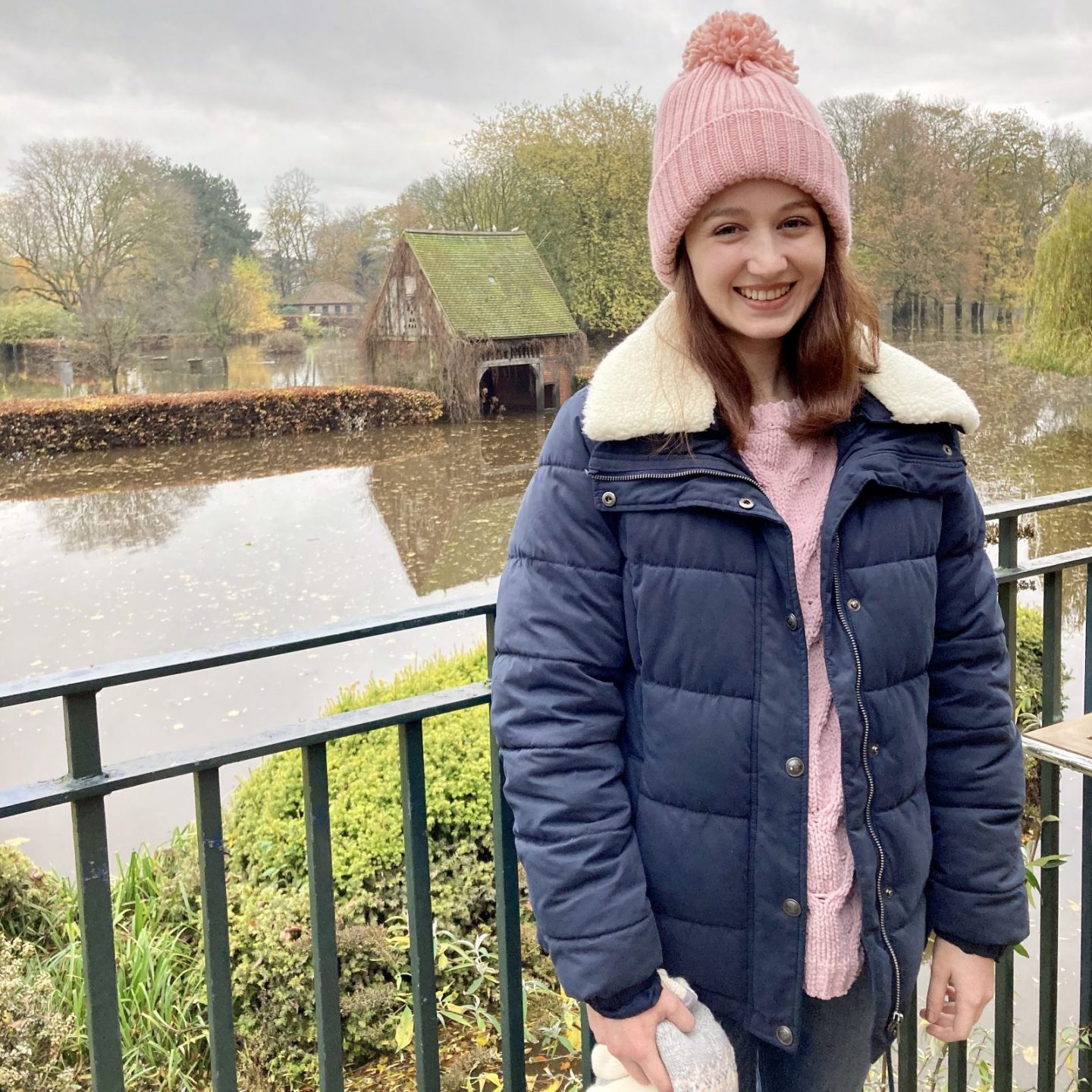 Pippa stood outdoors and smiling, wearing navy blue winter coat, lilac jumper and pale pink bobble hat. Park visible in back (rather flooded with water at the time!)