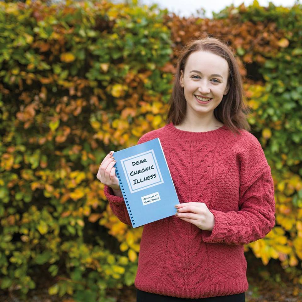 pippa stood outdoors wearing a red jumper, holding up a blue paperback copy of dear chronic illness - our charity book
