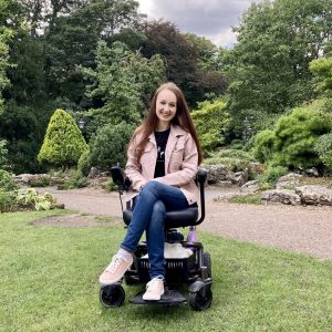 pippa outdoors in york museum gardens, surrounded by trees and grass. pippa is sat in small black electric wheelchair, smiling with hands in lap, wearing pale pink denim jacket and jeans.