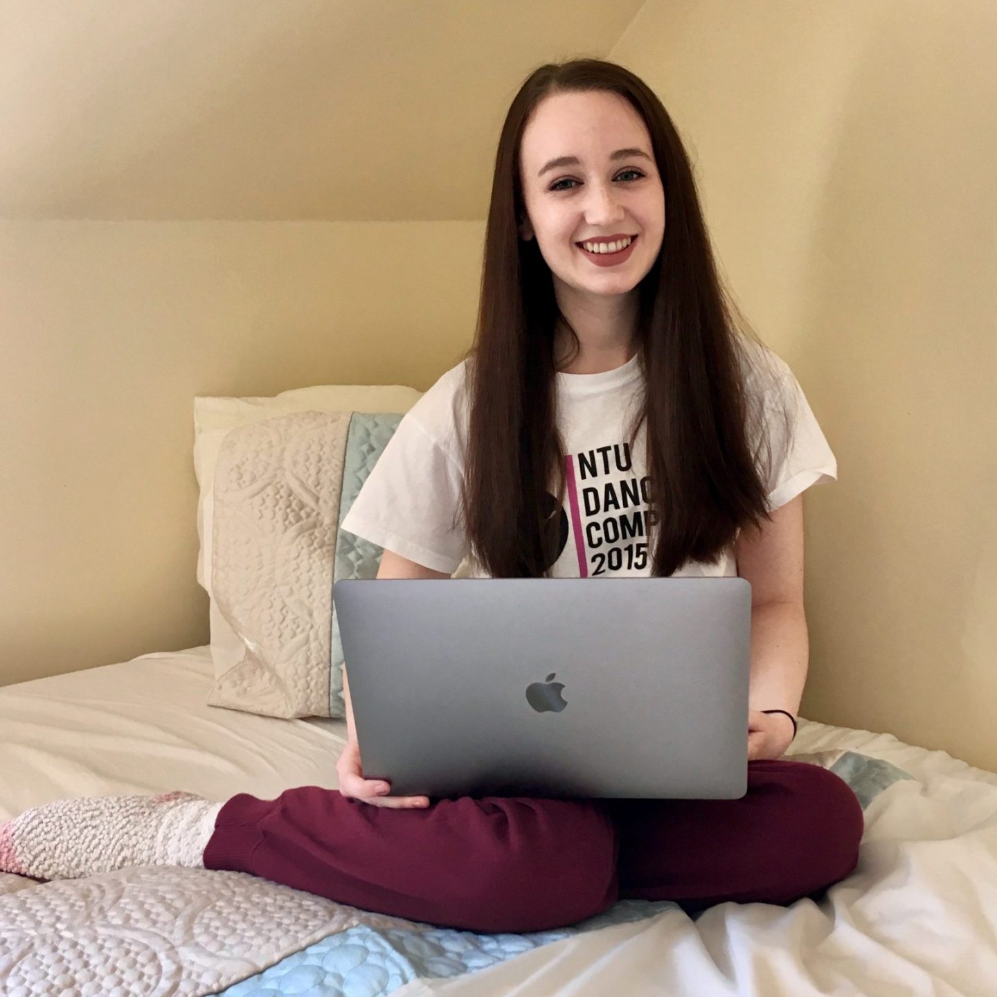 pippa sat on bed, wearing comfy clothes, with laptop propped up on her crossed legs. pippa has long brown hair down and is smiling 