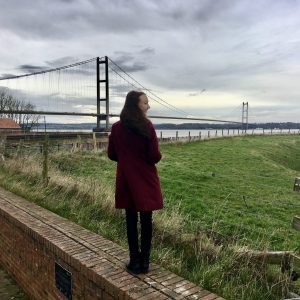 pippa stood on wall outdoors, back to camera and looking at the humber bridge in the distance. pippa is wearing a long burgandy coat with black jeans and boots