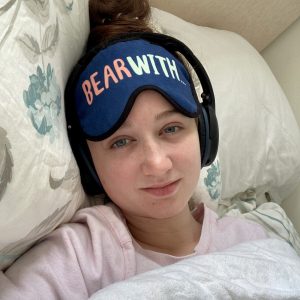 selfie of pippa in bed, no make-up and looking exhausted, wearing pink pyjamas, black noise cancelling headphones and a blue eyemask with the words 'bear with' printed on the front