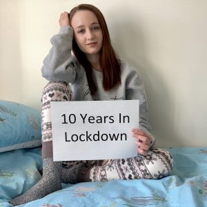 pippa sat on top of made bed, wearing pink and grey pyjamas, long brown hair down. pippa has one leg propped up with her hand resting on it, the other hand holding a sign which says '10 years in lockdown'