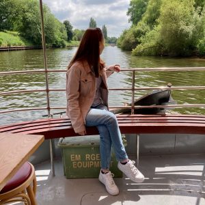 pippa sat on bench onboard boat that's out on the river, one hand on the rails and looking behind her. pippa is wearing a light pink denim jacket, blue jeans and white trainers.