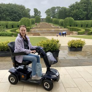 pippa sat in small black mobility scooter facing the side, at the alnwick garden with the grand cadcade waterfall feature in the background. pippa is wearing a lilac rain coat, jeans, and grey trainers, hair in two french plaits and smiling