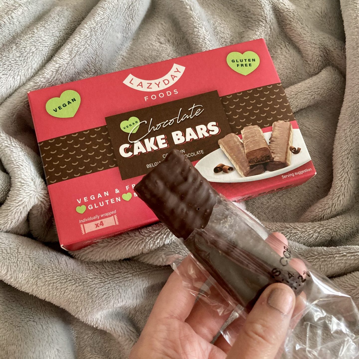 pippa's hand holding an unwrapped chocolate cake bar in her hand, with lazy day foods chocolate cake bar cardboard packaging in background