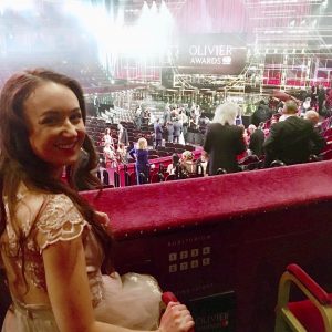 pippa sat in box at royal albert hall, looking back towards the camera. pippa is in a formal pink dress and olivier awards stage is visible in background