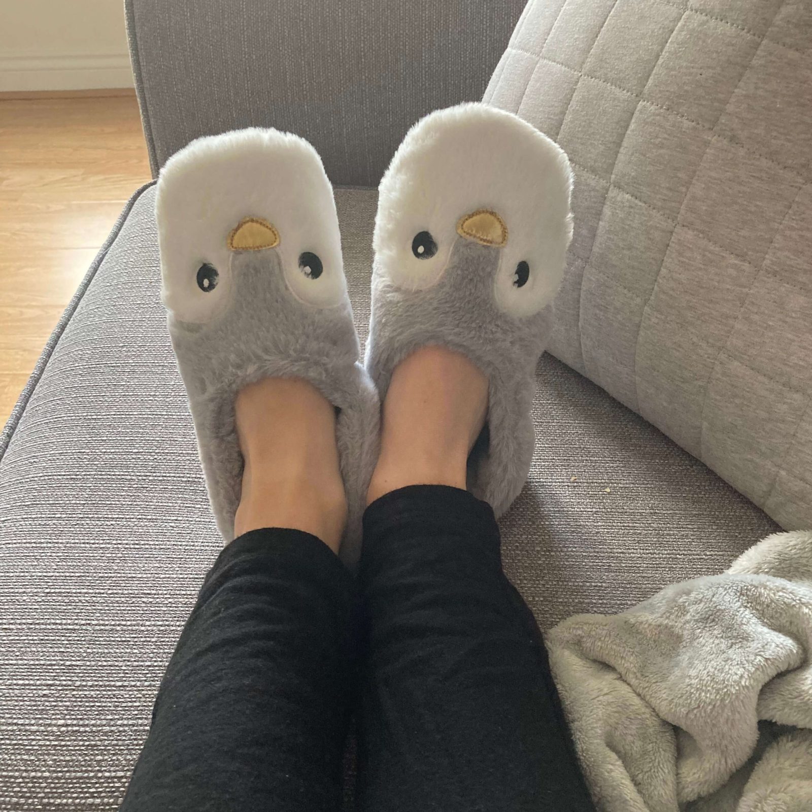 pippa's legs propped up on grey sofa with grey cushions in background. on pippa's feet are novelty penguin chick slippers, grey and white in design and with heating pads underneath